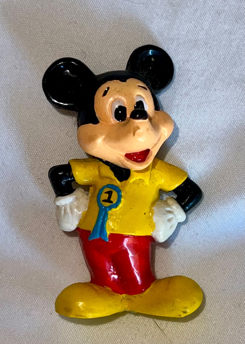 Peut inclure: Une figurine en plastique jaune et rouge de Mickey Mouse portant une chemise jaune avec un ruban bleu sur lequel est inscrit "1".