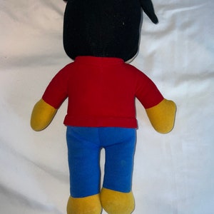 May include: A plush toy with black ears, a red shirt, blue pants, and yellow shoes. The toy is standing on a white surface.