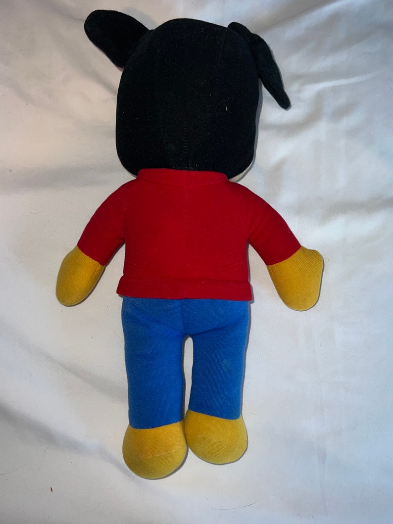 May include: A plush toy with black ears, a red shirt, blue pants, and yellow shoes. The toy is standing on a white surface.