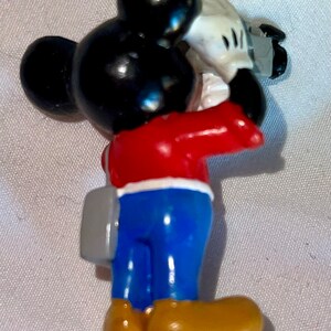 May include: A red, white, and blue Mickey Mouse figurine holding a camera. He is wearing blue pants and brown shoes. He has a gray bag slung over his shoulder.