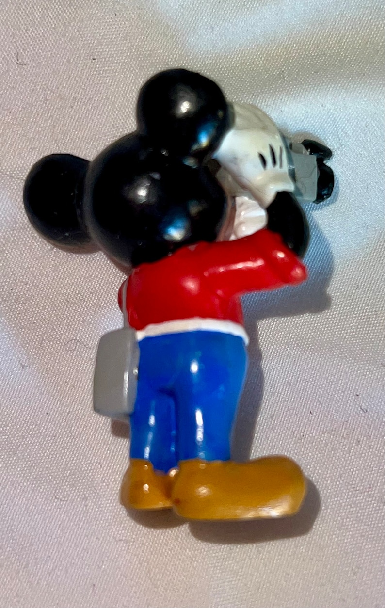 May include: A red, white, and blue Mickey Mouse figurine holding a camera. He is wearing blue pants and brown shoes. He has a gray bag slung over his shoulder.