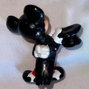 May include: A small, black and white figurine of a cartoon mouse in a tuxedo, holding a black top hat. The mouse has a red bow tie and white gloves. The figurine is a collectible item.