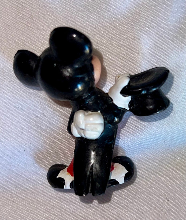 May include: A small, black and white figurine of a cartoon mouse in a tuxedo, holding a black top hat. The mouse has a red bow tie and white gloves. The figurine is a collectible item.