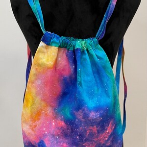 May include: A drawstring bag with adjustable straps, featuring a galaxy print in shades of blue, pink, purple, and yellow.
