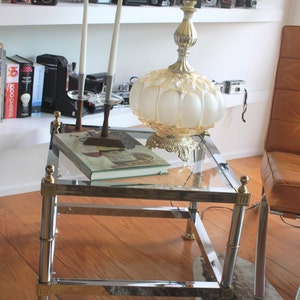 Puede incluir: Una mesa de centro de vidrio con marco cromado y detalles en latón. La mesa tiene dos niveles y está sobre un suelo de madera. Una lámpara con una pantalla de vidrio blanco esmerilado y una pantalla de tela beige está sobre la mesa. La lámpara está sobre un libro.