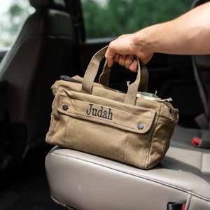 May include: A tan canvas tote bag with the name "Judah" embroidered in black. The bag has two handles, a front pocket with a snap closure, and is being held in a car.