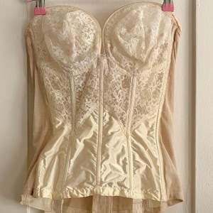May include: A cream-colored strapless corset featuring a floral lace overlay on the bust and a smooth satin midsection. The garment includes vertical boning, a sweetheart neckline, and four attached garter straps with metal clips at the hem.