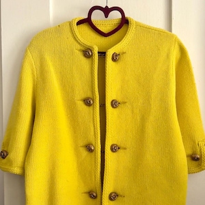 May include: A bright yellow knit cardigan featuring a round neckline, three-quarter length sleeves, and a front closure with four metallic gold buttons. Each sleeve cuff is also adorned with a single matching gold button, creating a cohesive look.
