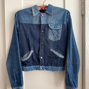 Könnte beinhalten: Vintage Jeansjacke in Blautönen. Sie hat ein zweifarbiges Design mit einem dunkelblauen Körper und hellblauen Ärmeln und Kragen. Die Jacke hat eine Knopfleiste, eine Brusttasche und zwei Vordertaschen. Die Jacke hängt an einem Kleiderbügel.