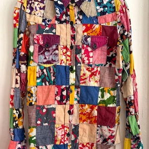 Op de afbeelding: Een patchworkshirt met lange mouwen, een knoopsluiting en twee borstzakken. Het shirt is samengesteld uit verschillende vierkanten stof in levendige kleuren en patronen, waaronder rood, blauw, geel en groen. De kraag is donkerpaars.