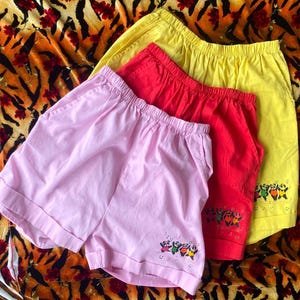 May include: Three pairs of cotton shorts with a playful panda design. The shorts are pink, red, and yellow.