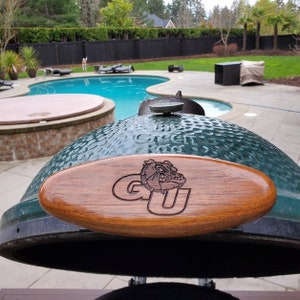 May include: A green ceramic grill with a wooden handle featuring the letters "GU" and a bulldog logo. The grill is outdoors near a pool.