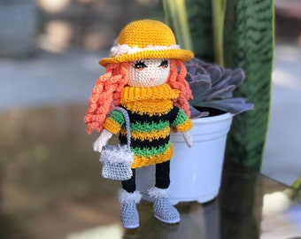 May include: A handmade crochet doll with orange curly hair, wearing a yellow hat, a black, yellow, and green striped sweater, black pants, and gray boots. The doll has a small gray purse and is standing in front of a plant.