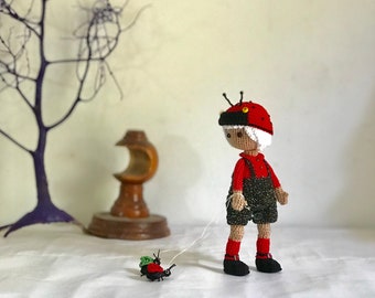 May include: A handmade crochet doll dressed as a ladybug, wearing a red shirt and overalls with black shorts. The doll has a red hat with black spots and antennae. A smaller ladybug is attached by a string.