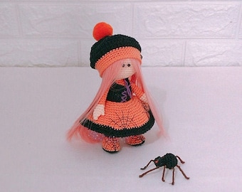 Puede incluir: Una muñeca de crochet que lleva un vestido negro y naranja con un diseño de telaraña. La muñeca tiene el pelo rosa y un sombrero negro con un pompón naranja. Una araña negra está sentada en la superficie blanca frente a la muñeca.