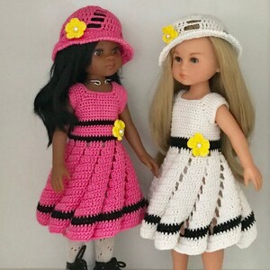 May include: Two dolls wearing crocheted dresses and hats. The doll on the left is wearing a pink dress with black trim and a pink hat with a yellow flower. The doll on the right is wearing a white dress with black trim and a white hat with a yellow flower.