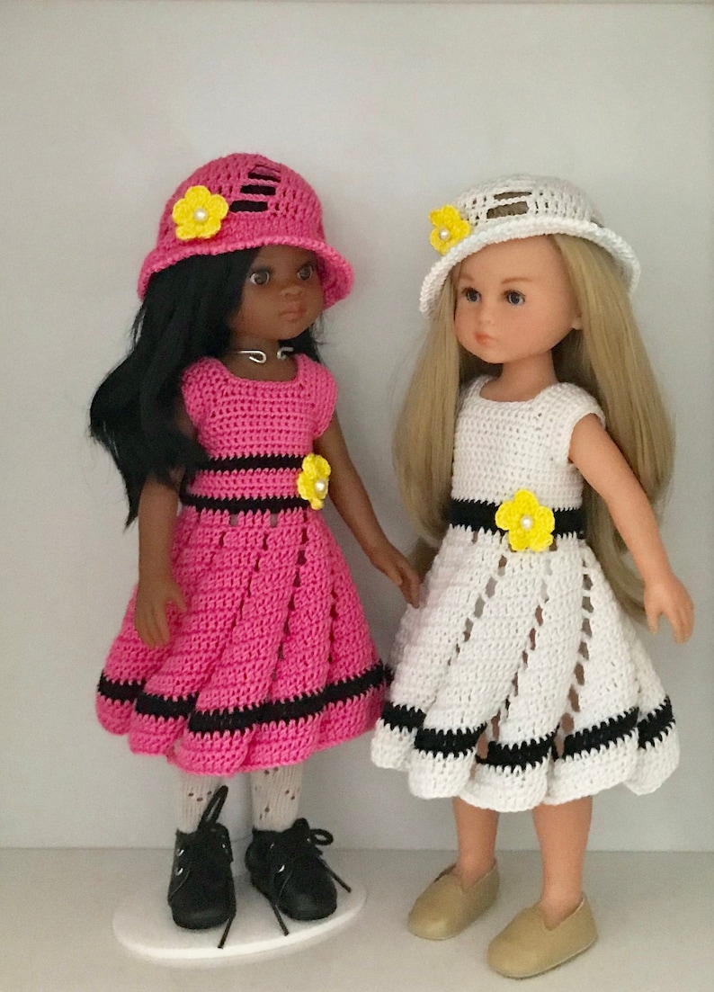 May include: Two dolls wearing crocheted dresses and hats. The doll on the left is wearing a pink dress with black trim and a pink hat with a yellow flower. The doll on the right is wearing a white dress with black trim and a white hat with a yellow flower.