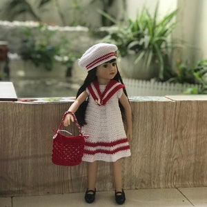 May include: A white crocheted dress with red stripes and a white crocheted sailor hat. The doll is holding a red crocheted handbag.
