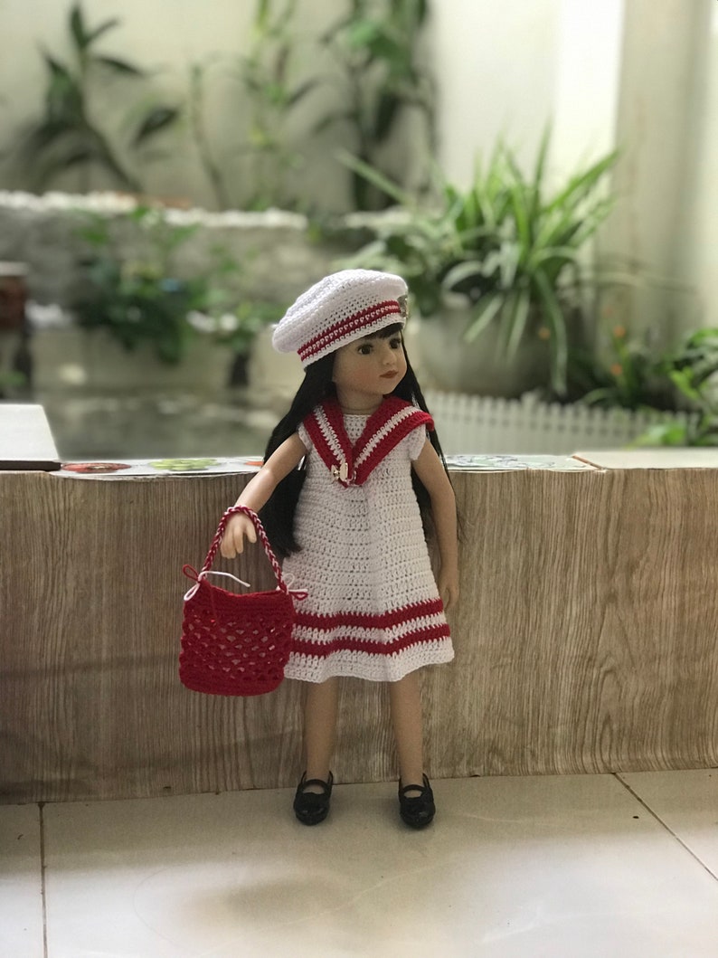 May include: A white crocheted dress with red stripes and a white crocheted sailor hat. The doll is holding a red crocheted handbag.