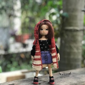 May include: A handmade crochet doll wearing a black top, denim skirt, and a red and cream striped hooded jacket. The doll has brown hair and is wearing black shoes with red accents. The doll is standing on a wooden surface.