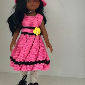 May include: A pink crocheted dress with black stripes and yellow flower accents. The doll is wearing a pink crocheted hat with black stripes and a yellow flower accent. The doll has black hair and brown eyes.