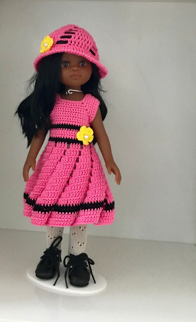 May include: A pink crocheted dress with black stripes and yellow flower accents. The doll is wearing a pink crocheted hat with black stripes and a yellow flower accent. The doll has black hair and brown eyes.