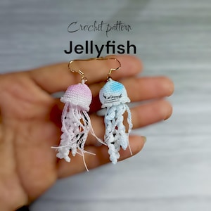 May include: Handmade crochet jellyfish earrings. One earring is pink and white, the other is blue and white. The earrings have gold-coloured ear wires and white, dangling tentacles. The text "Crochet pattern Jellyfish" is above the earrings.