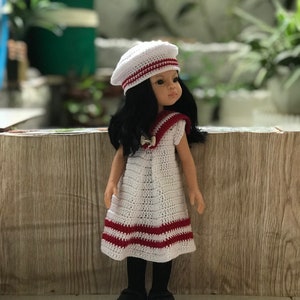 May include: A white crocheted dress with red stripes, worn by a doll with black hair and a white crocheted beret. The doll is standing in front of a wood-grain background.