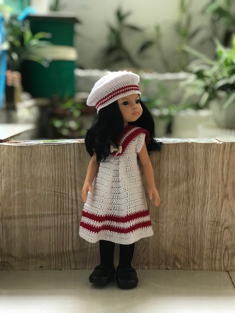 May include: A white crocheted dress with red stripes, worn by a doll with black hair and a white crocheted beret. The doll is standing in front of a wood-grain background.