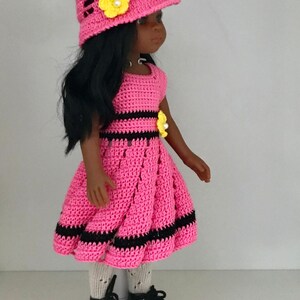 May include: A pink crocheted dress with black stripes and a yellow flower accent. The doll is wearing a pink crocheted hat with a yellow flower and black shoes.