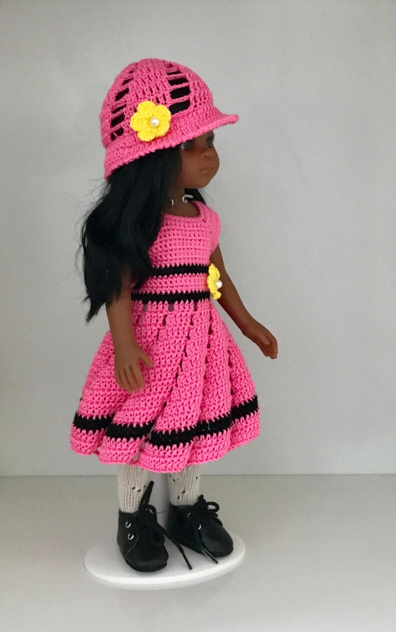 May include: A pink crocheted dress with black stripes and a yellow flower accent. The doll is wearing a pink crocheted hat with a yellow flower and black shoes.