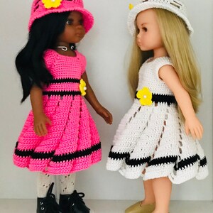 May include: Two dolls wearing crocheted dresses and hats. The doll on the left is wearing a pink dress with black trim and a pink hat with a yellow flower. The doll on the right is wearing a white dress with black trim and a white hat with a yellow flower.