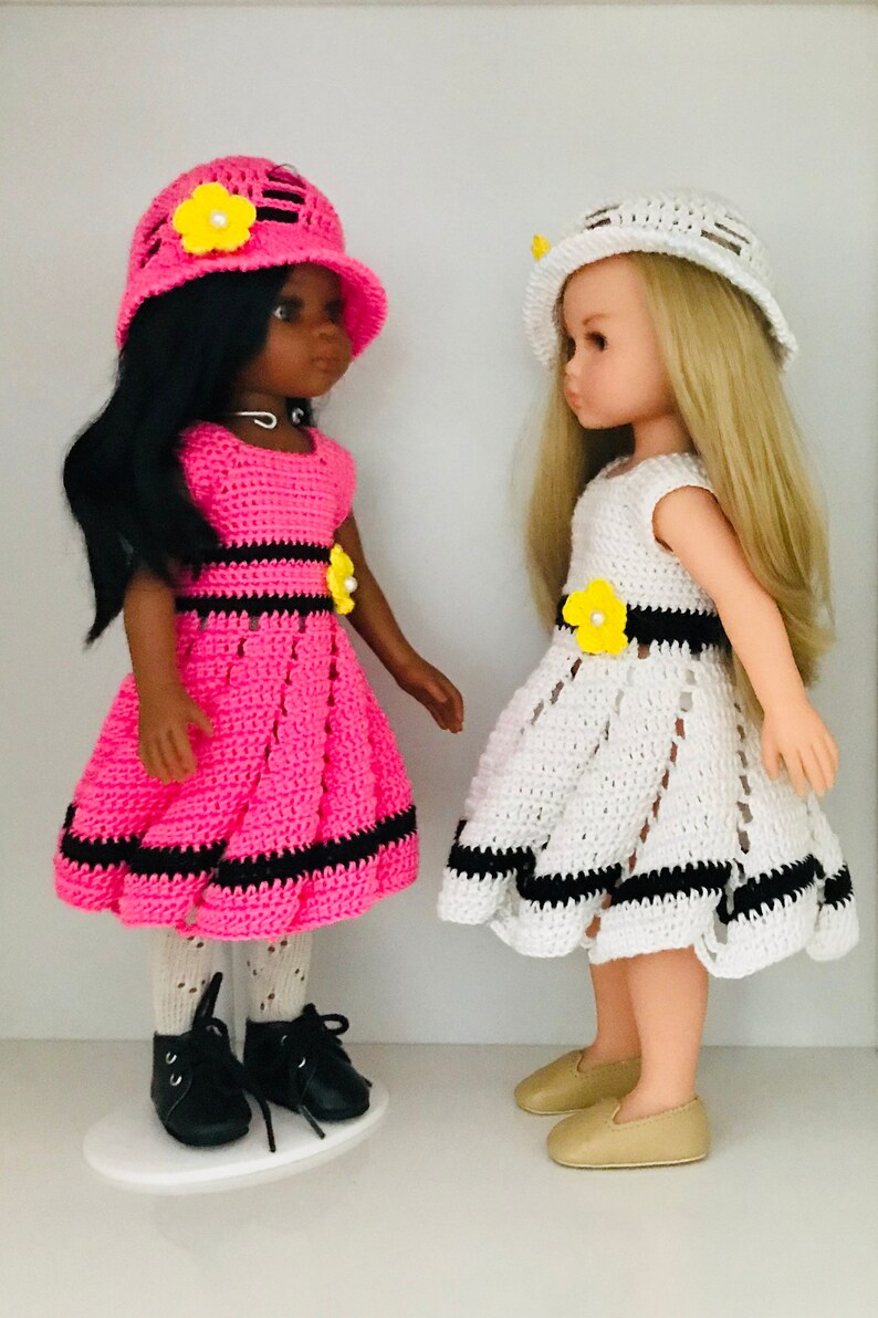 May include: Two dolls wearing crocheted dresses and hats. The doll on the left is wearing a pink dress with black trim and a pink hat with a yellow flower. The doll on the right is wearing a white dress with black trim and a white hat with a yellow flower.