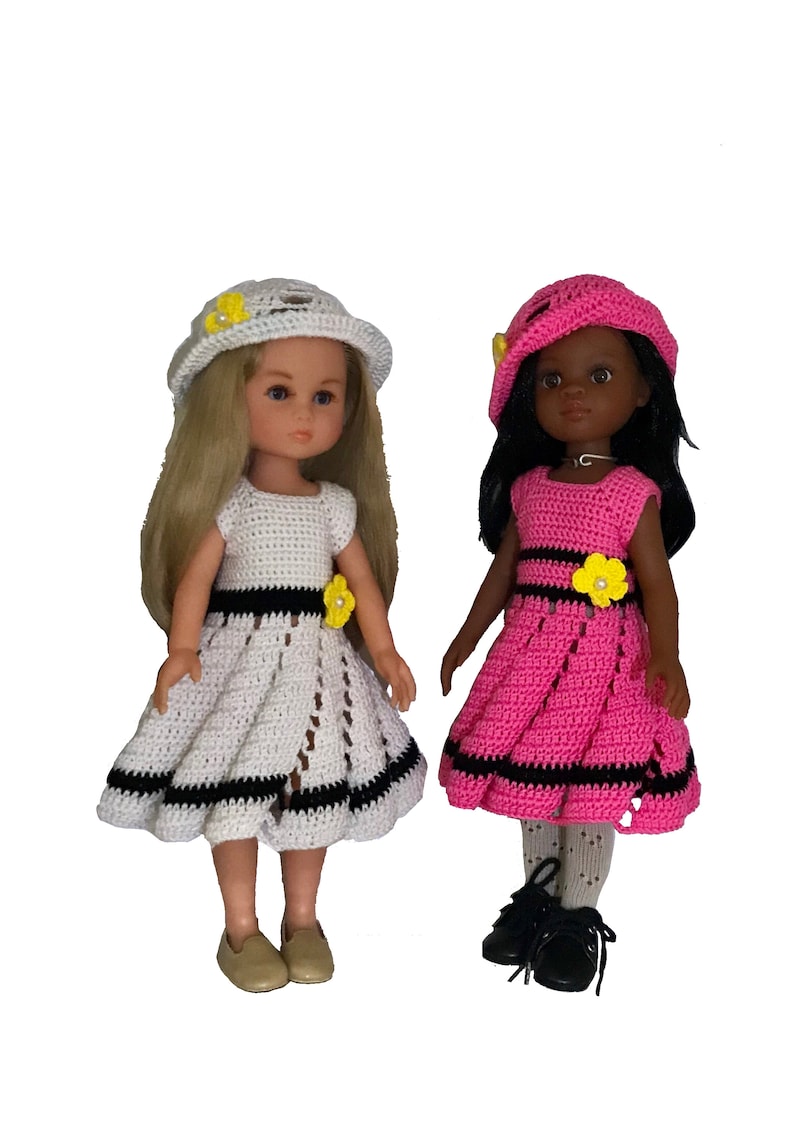 May include: Two dolls wearing crocheted dresses and hats. The doll on the left is wearing a white dress with black trim and a white hat with a yellow flower. The doll on the right is wearing a pink dress with black trim and a pink hat with a yellow flower.