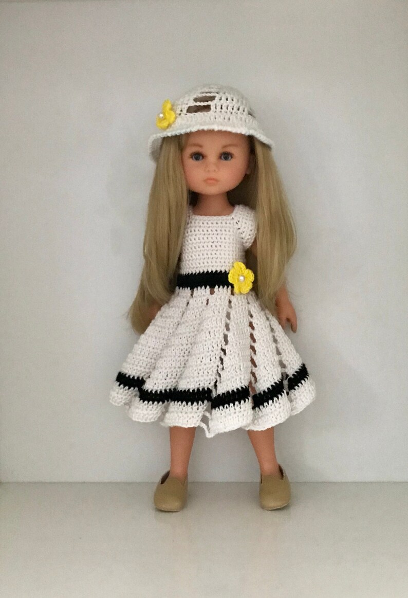 May include: A white crocheted dress with black stripes and yellow flower accents. The doll is wearing a white crocheted hat with yellow flowers and brown shoes.