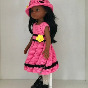 May include: A pink crocheted dress with black stripes and a yellow flower accent. The doll is wearing black shoes and a pink crocheted hat.