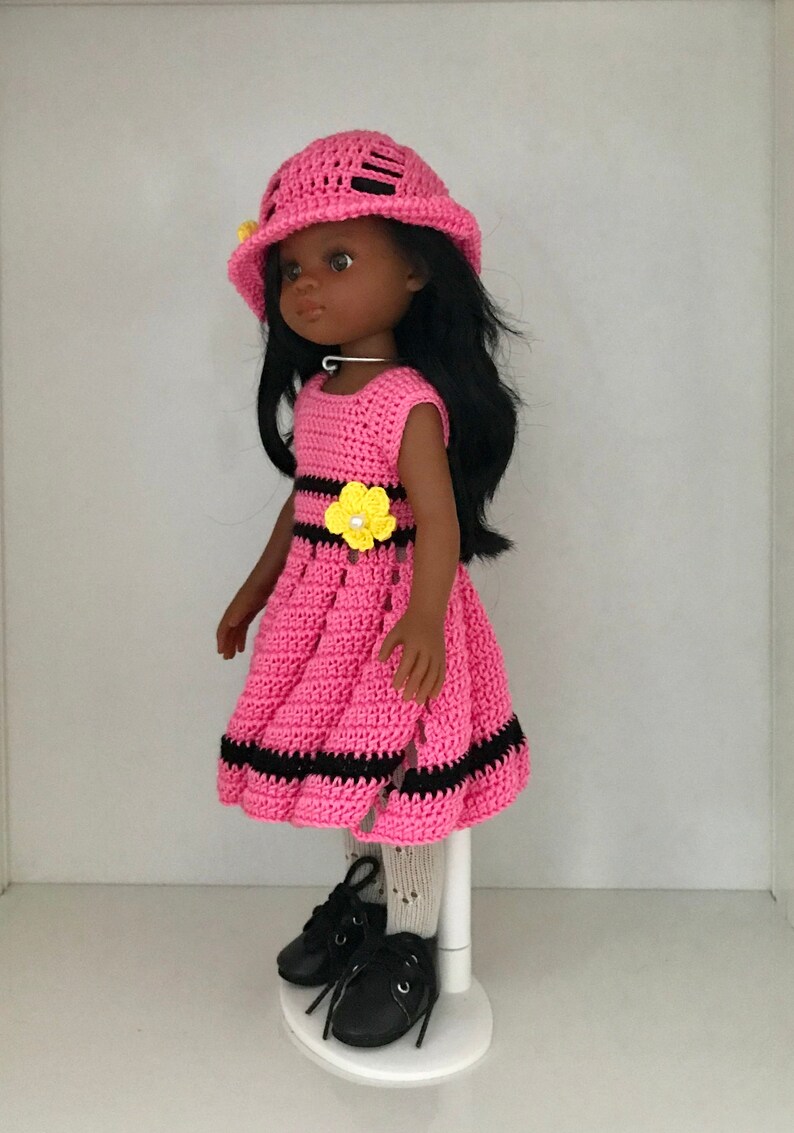 May include: A pink crocheted dress with black stripes and a yellow flower accent. The doll is wearing black shoes and a pink crocheted hat.