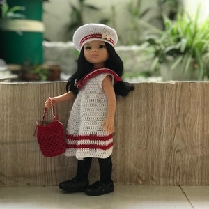 May include: A doll wearing a white crocheted dress with red stripes, a white sailor hat with a gold anchor, and black shoes. The doll is holding a red crocheted bag.