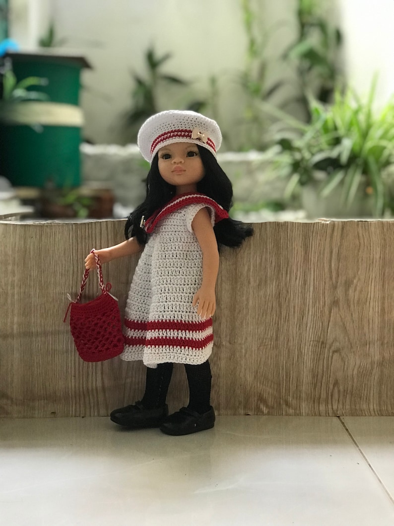 May include: A doll wearing a white crocheted dress with red stripes, a white sailor hat with a gold anchor, and black shoes. The doll is holding a red crocheted bag.