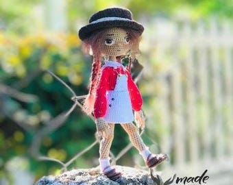 Puede incluir: Muñeca de ganchillo hecha a mano con sombrero marrón con banda blanca, chaqueta roja y vestido blanco con peto. La muñeca tiene pelo castaño y está de pie sobre una roca. Lleva zapatos marrones y calcetines blancos. El texto "made by amigurumi" está en la esquina inferior derecha.