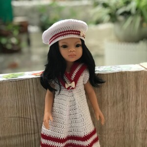May include: A white crocheted dress with red stripes and a sailor collar. The dress is worn by a doll with black hair and a white crocheted beret with a gold pin.