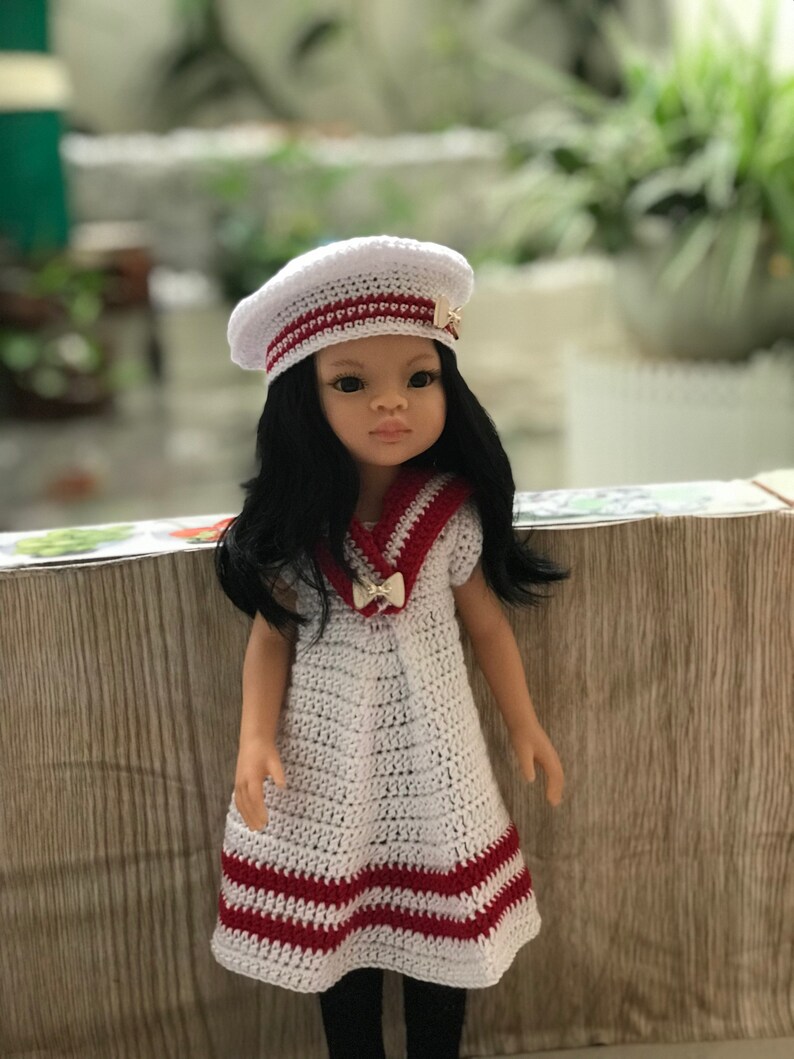 May include: A white crocheted dress with red stripes and a sailor collar. The dress is worn by a doll with black hair and a white crocheted beret with a gold pin.