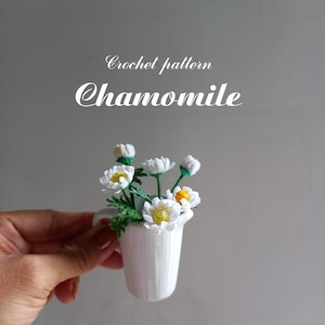 May include: Crochet pattern Chamomile: A close-up of a crocheted bouquet of white chamomile flowers with yellow centres in a small white vase.