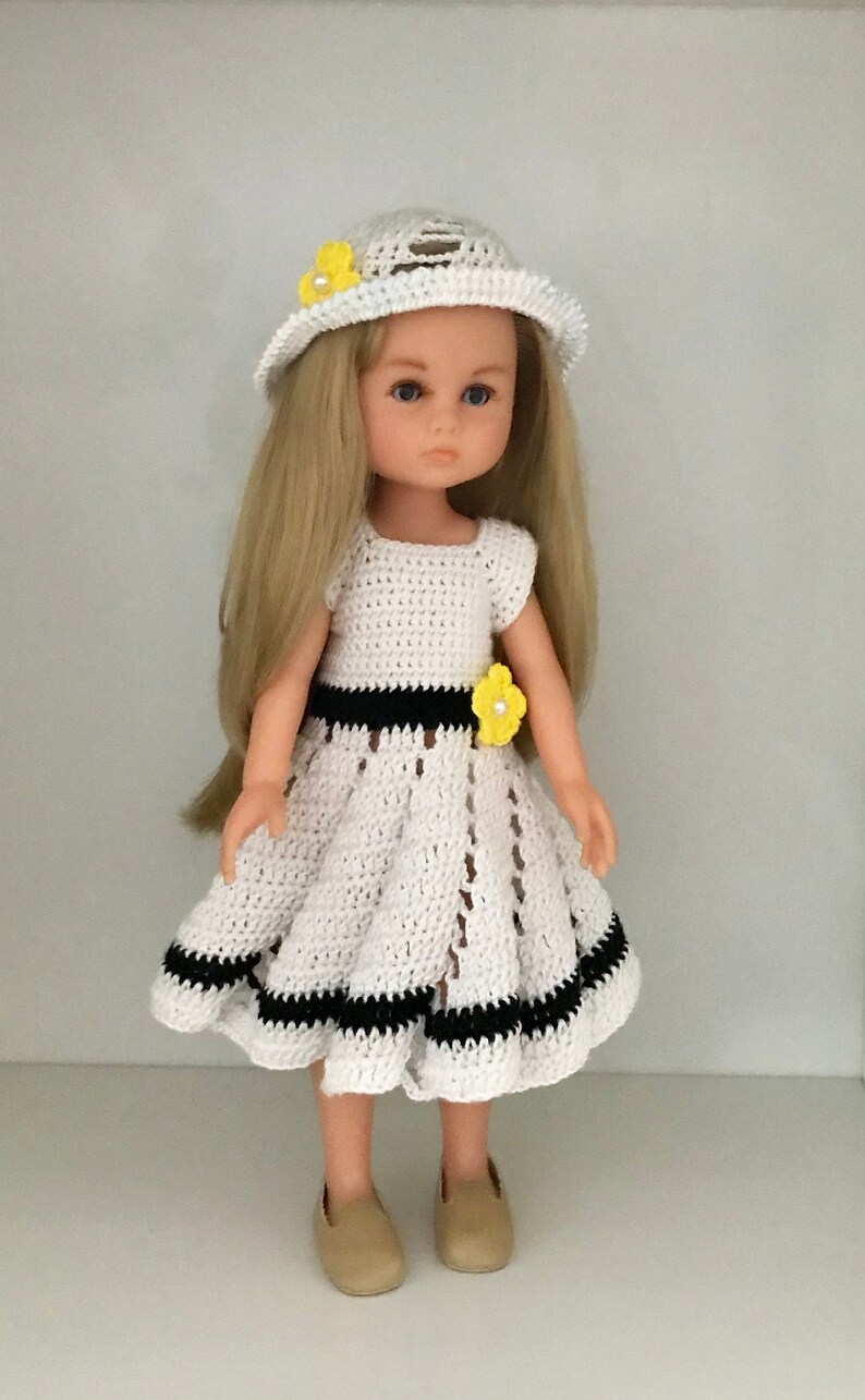 May include: A white crocheted dress with black trim and yellow flower accents. The dress is worn by a doll with blonde hair and a white crocheted hat with a yellow flower.