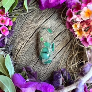 May include: An oval-shaped cabochon stone featuring a smooth, polished surface with a grey base color and distinct, rounded inclusions in varying shades of vibrant green, ranging from light lime to deep forest green, scattered across the face.