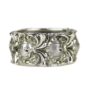 May include: A silver-colored ring with an ornate design. The ring features a repeating pattern of stylized floral and face-like elements. The details are finely crafted, with a textured surface and intricate relief work. The ring is set against a white background.