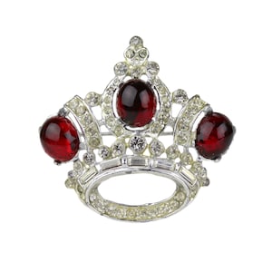 Könnte beinhalten: Silberfarbene Kronenbrosche mit drei großen, runden, roten Edelsteinen und zahlreichen kleineren, klaren Strasssteinen. Die Brosche hat ein klassisches Kronen-Design mit kreisförmiger Basis und spitz zulaufenden Bögen. Ein Schmuckstück im Vintage-Stil.