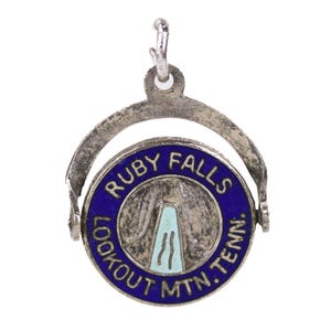 May include: A silver-toned charm with a rotating circular pendant. The pendant features a blue border with the words "RUBY FALLS LOOKOUT MTN. TENN." encircling a central image of a waterfall in a light blue color. The charm has a loop for attachment.