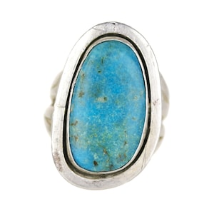 May include: A silver ring featuring a large, oval-shaped turquoise stone with natural brown matrix inclusions. The stone is set within a smooth, polished silver bezel and a wide, rounded silver band that frames the central turquoise gemstone.