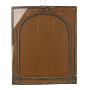 May include: A rectangular antique picture frame with a decorative metal border. The frame features an arched inner design and ornate corner and top accents. The background is a textured brown material, and the frame includes a glass pane.